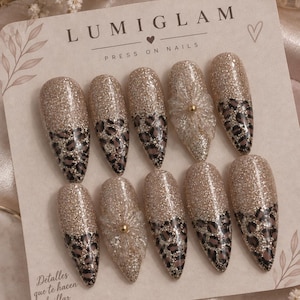 Puede incluir: Un juego de diez uñas postizas en forma de almendra sobre una tarjeta con la marca LUMIGLAM. Las uñas tienen un acabado de purpurina dorada combinado con un estampado de leopardo marrón y negro. Dos uñas llevan un adorno floral transparente.