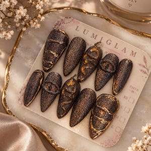 Puede incluir: Un juego de diez uñas postizas en forma de almendra con un acabado oscuro y brillante. Las uñas presentan diseños en relieve de serpientes y ojos en oro metálico. Se muestran en una tarjeta de la marca Lumiglam con tallas XS, S, M y L.