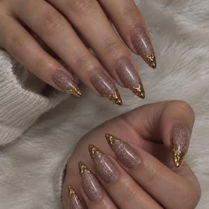 Puede incluir: Uñas en forma de almendra con una base de purpurina de color marrón claro brillante. Cada uña está acentuada con una punta metálica dorada texturizada que imita el aspecto de metal fundido, creando un contraste con la base neutra brillante.