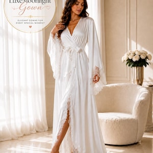 Op de afbeelding: Een kamerjas van wit satijn tot op de grond met lange, wijde mouwen met kanten afwerking, een V-hals en een bijpassende ceintuur. De tekst LuxeMoonlight Gown Elegant Gowns For Every Special Moment is zichtbaar in een rond logo.