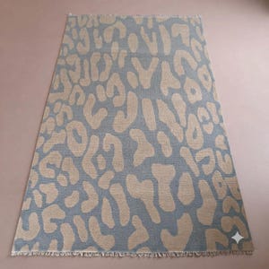 May include: A rectangular area rug featuring a repeating leopard print pattern in shades of slate blue and beige. The rug has a woven texture and is finished with short, frayed fringe detailing along the top and bottom edges of the fabric.