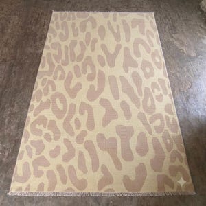 May include: A rectangular area rug featuring a repeating leopard print pattern in shades of cream and light tan. The rug has a woven texture and is finished with short, straight fringe along the top and bottom edges, creating a clean look.