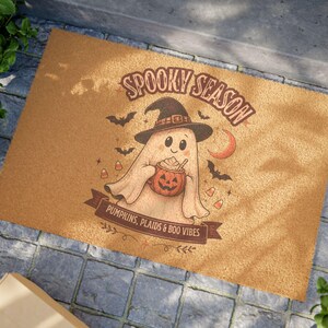 May include: A rectangular tan coir doormat featuring a brown graphic of a ghost wearing a witch hat, holding a pumpkin mug. The text reads SPOOKY SEASON and PUMPKINS, PLAIDS & BOO VIBES, surrounded by small bats, a crescent moon, and candy corn icons.