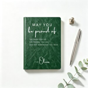 Pode incluir: Um diário verde-escuro com capa texturizada apresentando o texto em branco: MAY YOU be proud of THE WORK YOU DO THE PERSON YOU ARE AND THE DIFFERENCE YOU MAKE FOR Olivia. Uma caneta prateada e um pequeno objeto de metal prateado estão próximos.