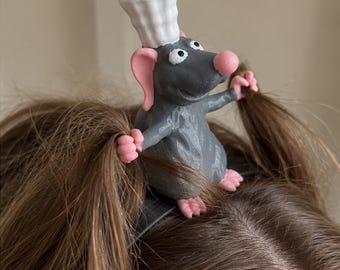 May include: A dark gray rat figurine wearing a white chef hat, attached to a black hair clip. The rat has pink ears, a pink nose, and pink paws that are positioned to hold two sections of brown hair on either side of the hair accessory.