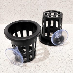 May include: Two black plastic cylindrical net pots with grid-like walls and wide rims. Each pot features a clear, circular suction cup attached to the side, designed for mounting the containers to smooth surfaces for aquatic or plant display.