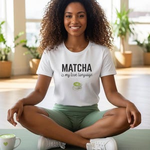 May include: A white short-sleeved t-shirt featuring the black text "MATCHA is my love language" and a small graphic of a green matcha latte in a cup. The shirt is paired with sage green athletic shorts and white sneakers.