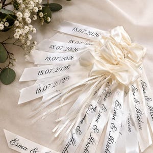 May include: A collection of off-white satin ribbons tied into bows, each printed with the date 18.07.2026 and the names Esma & Ibrahim in black script. The ribbons are arranged in a fan shape, featuring smooth fabric texture and elegant lettering.