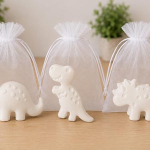 Puede incluir: Tres figuras de dinosaurios blancas hechas de un material liso y mate, cada una colocada frente a una pequeña bolsa de regalo de organza blanca transparente con cordón. Los dinosaurios presentan formas simples y redondeadas con patrones circulares.