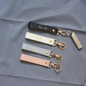 May include: Four rectangular leather keychains in black, cream, light blue, and light pink, each featuring gold-tone hardware and a gold-tone metal ring. The black keychain is embossed with No.98, while the others feature gold-tone lettering.