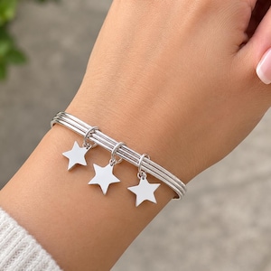 Peut inclure: Un bracelet jonc argenté orné de trois breloques en forme d'étoile. Le bracelet est composé de trois fines bandes métalliques parallèles qui entourent le poignet, chaque breloque étoile étant fixée par un petit anneau circulaire.