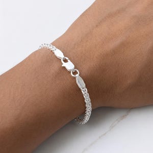 Puede incluir: Una pulsera de cadena de trigo plateada con acabado pulido y un cierre de mosquetón seguro. La pulsera está construida con eslabones metálicos entrelazados que crean una apariencia texturizada y redondeada, rodeando la muñeca con un diseño uniforme.