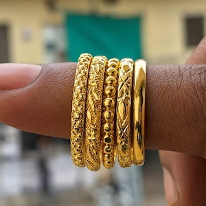 May include: A set of five gold rings featuring distinct textures, including a beaded band, a floral engraved band, a diamond-cut pattern, a textured stippled band, and one smooth polished gold band, all displayed together in a row.
