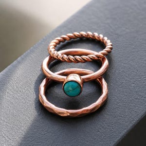 May include: A set of three copper-toned rings stacked together. The top ring features a twisted rope texture, the middle ring is a smooth band set with a round, polished turquoise stone, and the bottom ring has a hammered, irregular surface finish.
