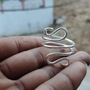 Puede incluir: Un anillo de color plateado con un diseño de alambre continuo que se enrolla alrededor del dedo. La banda metálica está formada por dos bucles redondeados distintos en la parte superior e inferior, creando un patrón abierto y giratorio pulido.