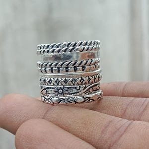 Könnte beinhalten: Ein Stapel aus sechs silberfarbenen Ringen mit verschiedenen Texturen und Mustern, darunter gedrehte Kordeldesigns, ein glattes Band, ein Perlenband, ein X-Muster, ein floral graviertes Band und ein Band mit Rautenmuster.