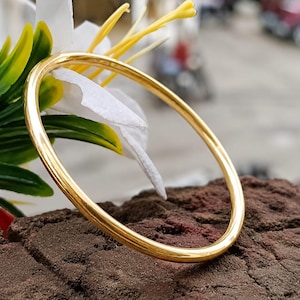 May include: A polished gold-tone bangle bracelet with a smooth, rounded surface. The circular metal band features a reflective finish that catches the light, showcasing a simple, continuous design without any clasps, hinges, or additional embellishments.