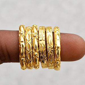 Può includere: Un set di sei anelli in metallo color oro, ognuno caratterizzato da un distinto motivo in rilievo. Gli anelli sono disposti fianco a fianco, mostrando varie texture tra cui forme geometriche, motivi floreali e superfici lisce e lucide.