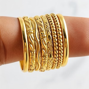 May include: A set of seven gold-toned rings worn together on a finger. The collection includes two plain polished bands on the ends, two bands with a beaded texture, and three central bands featuring intricate engraved patterns and wavy lines.