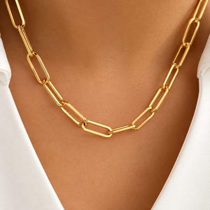 May include: A gold-toned chain necklace featuring elongated, rectangular paperclip-style links. The necklace has a polished, reflective finish and rests against a plain white collared shirt, showcasing a uniform, repeating link design throughout.