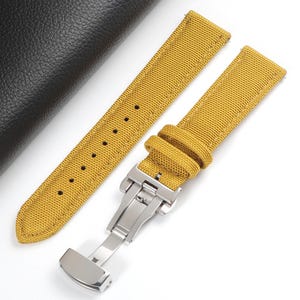 May include: A mustard yellow fabric watch strap featuring a textured weave, tonal stitching along the edges, and a silver-toned stainless steel deployment clasp. The strap includes two matching fabric keepers and a series of punched adjustment holes.
