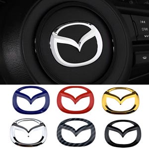 Può includere: Una collezione di sei emblemi per veicoli Mazda in varie finiture, tra cui blu, rosso, oro, cromo, motivo in fibra di carbonio e nero, mostrati sotto una vista ravvicinata di un logo Mazda argentato al centro di un volante nero.