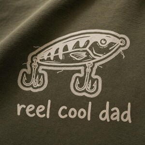 Puede incluir: Una prenda de tela verde oliva con un gráfico serigrafiado en blanco de un señuelo de pesca con dos anzuelos triples. Debajo del señuelo, el texto "reel cool dad" está impreso en una fuente blanca sencilla y en minúsculas.
