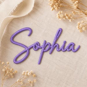 May include: A purple embroidered patch featuring the name Sophia in a cursive script font. The lettering has a raised, stitched texture and is centered on a light beige fabric background with small dried white flowers visible at the corners.