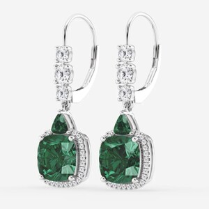 May include: A pair of silver drop earrings featuring a large cushion-cut dark green gemstone surrounded by a halo of small white stones. Above the main stone, a triangular green gem sits below three vertically aligned white stones on the hook.
