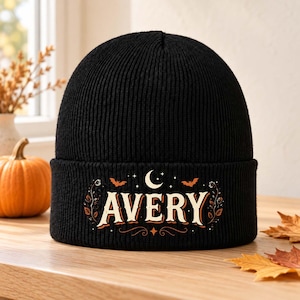 Könnte beinhalten: Eine schwarze, gerippte Strickmütze mit einem Grafikdruck auf dem Umschlag. Das Design zeigt den Namen AVERY in weißen Serifenschriftzeichen, umgeben von orangefarbenen und weißen Illustrationen eines Halbmonds, Sternen, Fledermäusen und Ranken.