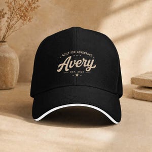 Könnte beinhalten: Eine schwarze Baseballkappe mit weißem Rand am Schirm. Die Vorderseite zeigt eine beige Grafik mit dem Text "BUILT FOR ADVENTURE", "Avery" und "EST. 2027", zentriert zwischen zwei kleinen Sternen und horizontalen Linien.
