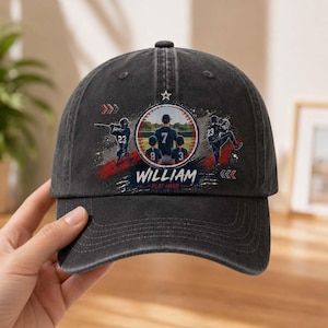 Puede incluir: Una gorra de béisbol gris carbón con un gráfico circular que muestra a tres jugadores de béisbol, el nombre WILLIAM y el texto PLAY HARD. El diseño incluye detalles en rojo y blanco, una pequeña estrella y líneas de movimiento.