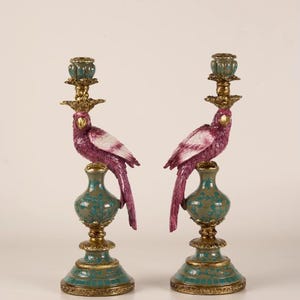 May include: A pair of candle holders featuring magenta and white parrots perched on olive green and teal ceramic bases with gold-toned metal accents. Each holder includes a decorative gold-toned metal top piece for a candle, supported by a parrot figure.