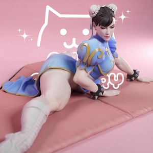 Puede incluir: Una figura de un personaje con cabello oscuro en moños, vistiendo un vestido azul con ribetes dorados, botas blancas y muñequeras negras con púas. El personaje está en posición de rodillas. Se ve un dibujo lineal blanco de una cara de gato.