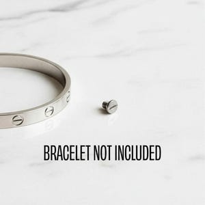 May include: A silver-colored bracelet with screw details and a single screw. The text "BRACELET NOT INCLUDED" is displayed in black. The bracelet is on a white marble surface.