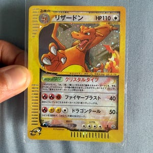 Puede incluir: Una carta coleccionable japonesa de Charizard con borde amarillo, que presenta una ilustración de la criatura naranja tipo dragón con llamas. La carta muestra el nombre リザードン, HP110 y texto en japonés que detalla ataques y estadísticas, en una funda protectora.
