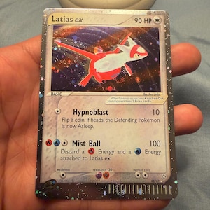 Puede incluir: Una carta coleccionable Latias ex con 90 HP, que presenta un fondo holográfico con temática espacial y estrellas. La carta muestra los ataques Hypnoblast y Mist Ball, junto con texto del juego, símbolos de energía y la fecha de copyright de 2003.