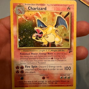 Könnte beinhalten: Eine holografische Charizard-Sammelkarte mit dem orangefarbenen, drachenähnlichen Wesen mit blauen Flügeln. Die Karte zeigt den Text Charizard, Stage 2, Flame Pokémon, Energy Burn und Fire Spin sowie einen Schadenswert von 100 Punkten.
