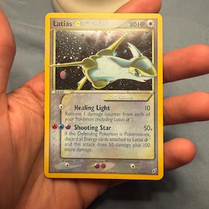Puede incluir: Una carta coleccionable Latias Star con 80 HP, con un borde amarillo y una ilustración de la criatura en el espacio. El texto de la carta incluye los ataques Healing Light y Shooting Star, con fecha de copyright de 2005 y número 105/107.