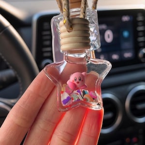 May include: A star-shaped glass bottle car air freshener hanging from a braided cord. The clear bottle contains a small pink Kirby figure surrounded by colorful, multi-shaped confetti pieces. It features a light-colored wooden cap and a clear glass top.
