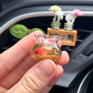May include: A miniature clear glass fishbowl containing a small orange goldfish figurine, light brown sand, and green aquatic plant accents. The bowl has a ruffled top edge and is held in a hand, showcasing its tiny scale and detailed design.