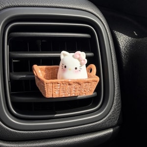 May include: A car air freshener clip featuring a white cat character with a pink bow sitting inside a small, light brown, rectangular woven basket. The character has black eyes, whiskers, and pink cheeks, attached to a black vehicle air vent.