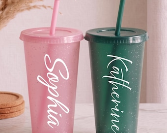 Puede incluir: Dos vasos de plástico reutilizables con tapas y pajitas. Un vaso es rosa translúcido con purpurina blanca y el nombre Sophia en letra blanca. El otro es verde oscuro translúcido con purpurina blanca y el nombre Katherine en letra blanca.