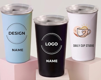 Puede incluir: Tres vasos de acero inoxidable con tapas transparentes. El de la izquierda es azul claro con un diseño circular y el texto DESIGN NAME. El central es negro con un diseño circular y el texto LOGO NAME. El de la derecha es blanco con un gráfico de taza de café y el texto DAILY CUP STUDIO.