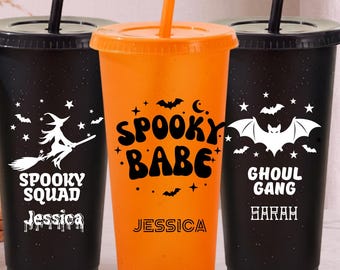 Puede incluir: Tres vasos de plástico con tapas y pajitas. Dos son negros con gráficos blancos de una bruja, estrellas, murciélagos y el texto "SPOOKY SQUAD JESSICA" y "GHOUL GANG SARAH". El vaso central es naranja con el texto negro "SPOOKY BABE JESSICA".