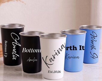 Puede incluir: Cinco vasos de acero inoxidable en negro, blanco y azul claro. Los vasos negros presentan el texto Amelia y Bottoms Up. El vaso blanco muestra el nombre Karina y Est.2026. Los vasos azul claro muestran el texto Worth It y Karina.