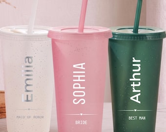 Puede incluir: Tres vasos de plástico con purpurina, tapas y pajitas en blanco, rosa y verde oscuro. Cada vaso tiene un nombre en texto blanco: Emilia, Sophia y Arthur. Debajo de los nombres aparecen los títulos Maid of Honor, Bride y Best Man con un corazón.