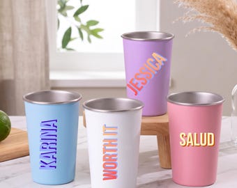 Puede incluir: Cuatro vasos de acero inoxidable con bordes metálicos en azul claro, blanco, lavanda y rosa. El vaso azul presenta el texto KARINA, el blanco dice WORTH IT, el lavanda muestra JESSICA y el rosa tiene la inscripción SALUD.