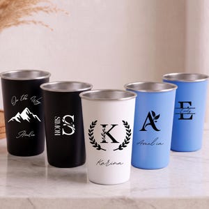 Puede incluir: Cinco vasos de acero inoxidable en negro, blanco y azul claro con bordes plateados. Los vasos negros presentan un gráfico de montaña con el texto On the Way Amelia y una letra S grande con el nombre Simon. El vaso blanco tiene una letra K dentro de una corona de laurel con el nombre Karina. Los vasos azules muestran una letra A con una hoja y el nombre Amelia, y una letra E con el nombre Emily.