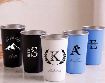 Puede incluir: Cinco vasos de acero inoxidable en negro, blanco y azul claro con bordes plateados. Los vasos negros presentan un gráfico de montaña con el texto On the Way Amelia y una letra S grande con el nombre Simon. El vaso blanco tiene una letra K dentro de una corona de laurel con el nombre Karina. Los vasos azules muestran una letra A con una hoja y el nombre Amelia, y una letra E con el nombre Emily.